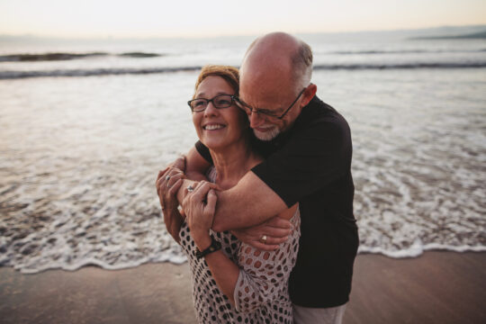 Retired Couple Cuddle Together Outside On Ocean Beach